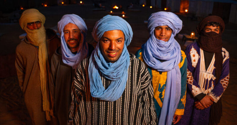 Nomadic Desert Team standing in the middle of their desert camp at night time illuminated by a fire.